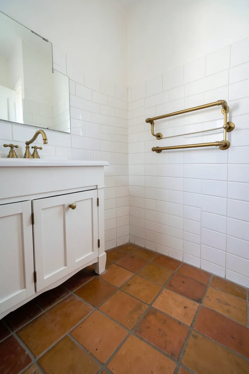 Hyper-realistic eye-level photograph of a Moroccan-style bathroom floor showing terracotta ceramic tiles with subtle variation and handmade quality, white subway tile walls with white grout, white vanity with brass faucet, brass towel rail on wall. Natural light. Materials: handmade terracotta ceramic tiles, white subway tiles, brass fixtures, white wood. Warm earthy terracotta floor. Authentic handmade tile details. No text, no logos, no watermarks.</p>
